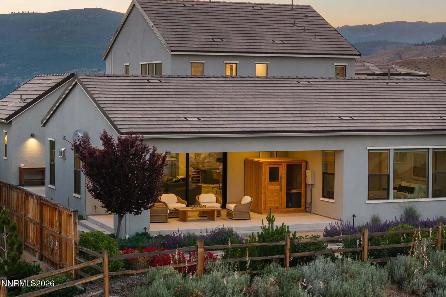 Modern gray home with a covered patio, outdoor seating, a wooden sauna, and a fenced garden against mountain views.