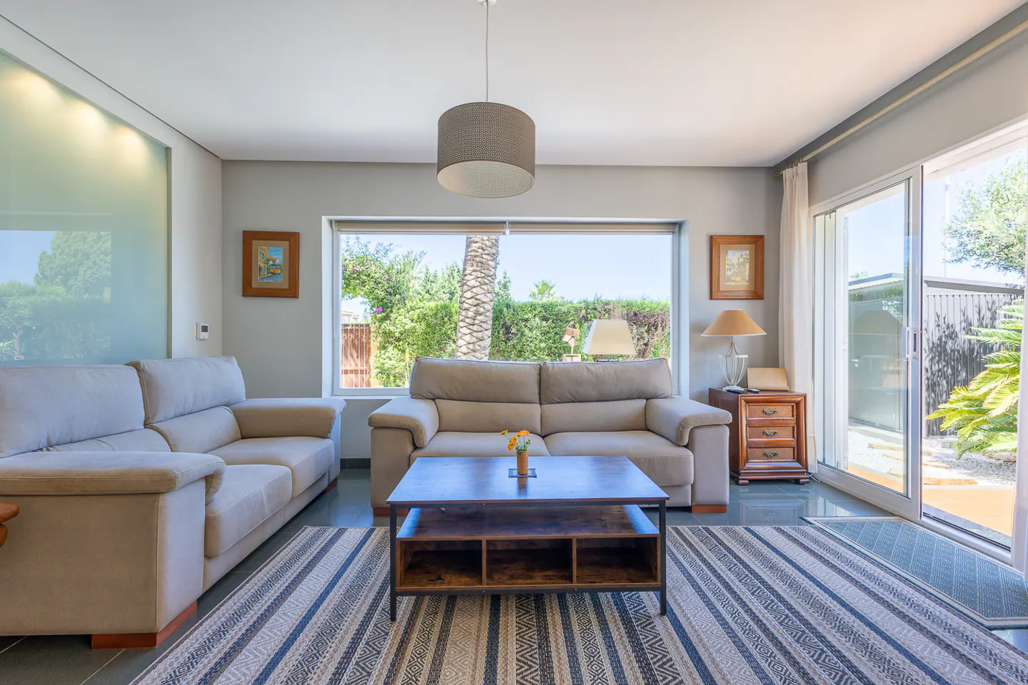 Living room with two beige sofas, a dark wood coffee table, and a striped rug. A large window overlooks a green garden.