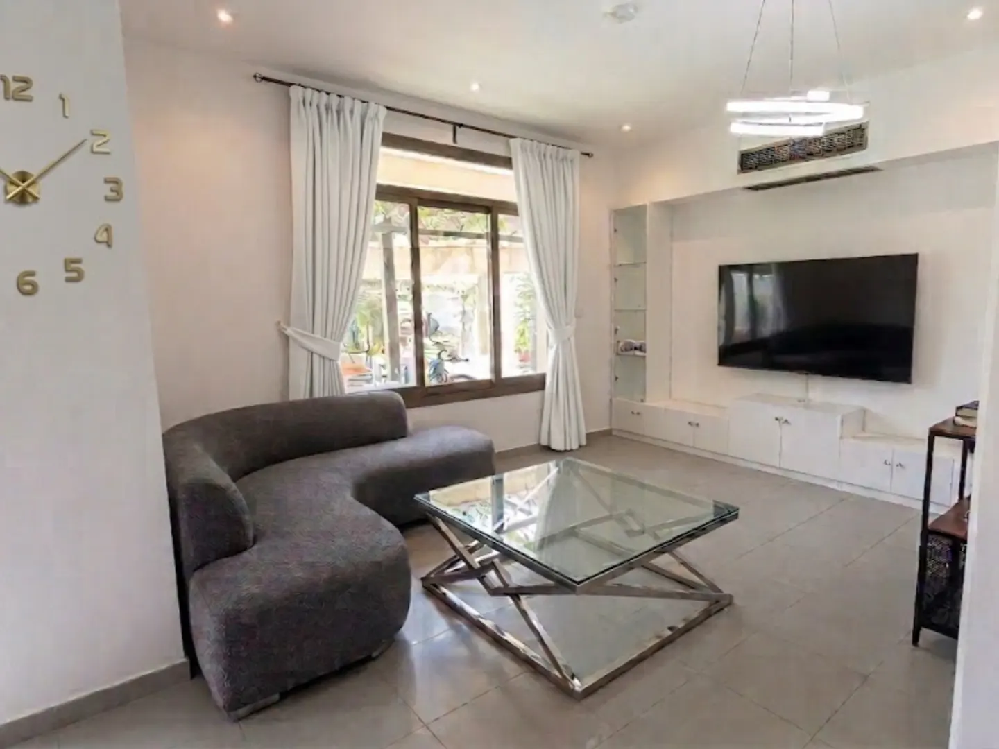 A bright living room with a gray curved sofa, glass coffee table, and a large TV on a white wall unit. A gold clock is on the wall.