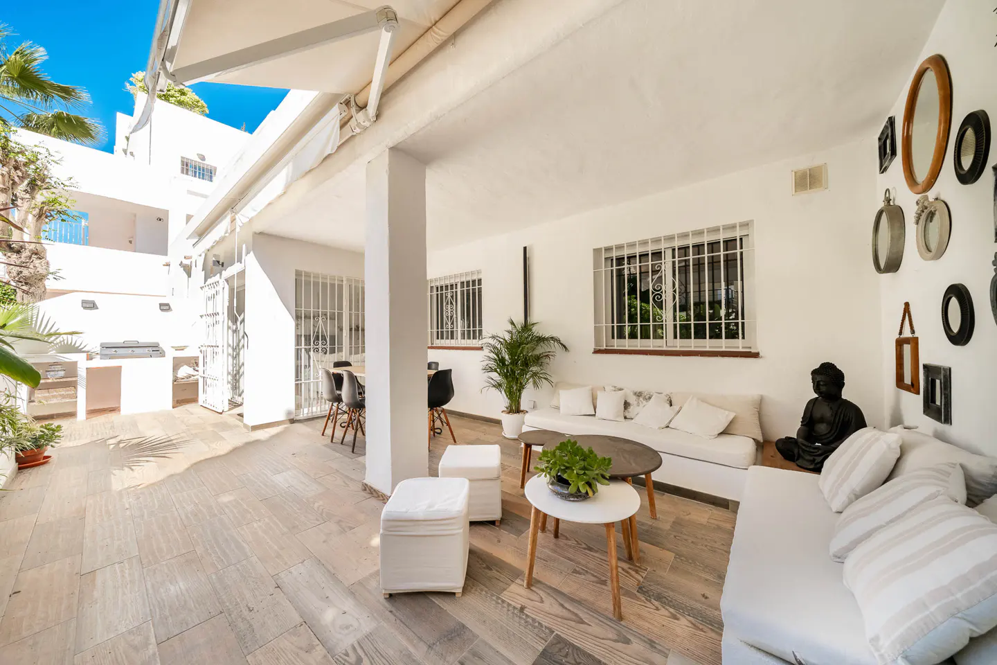 Outdoor patio with white sofas, tables, and a Buddha statue. A dining area and grill are visible in the background.
