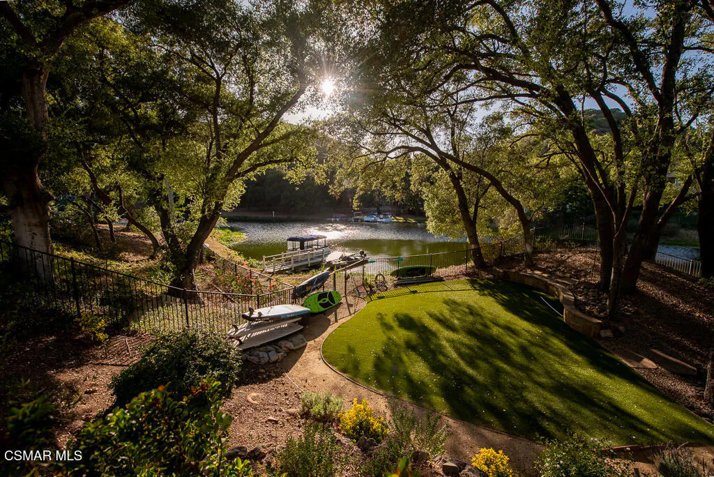 Lake view with dock, boats, and kayaks. Green lawn and trees surround the water. Sun shines through the trees.