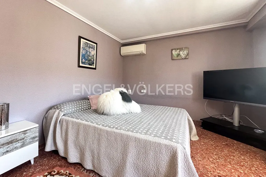Bedroom with a bed covered in a gray star-patterned quilt, a white bedside table, and a large TV on a stand. The walls are light purple.