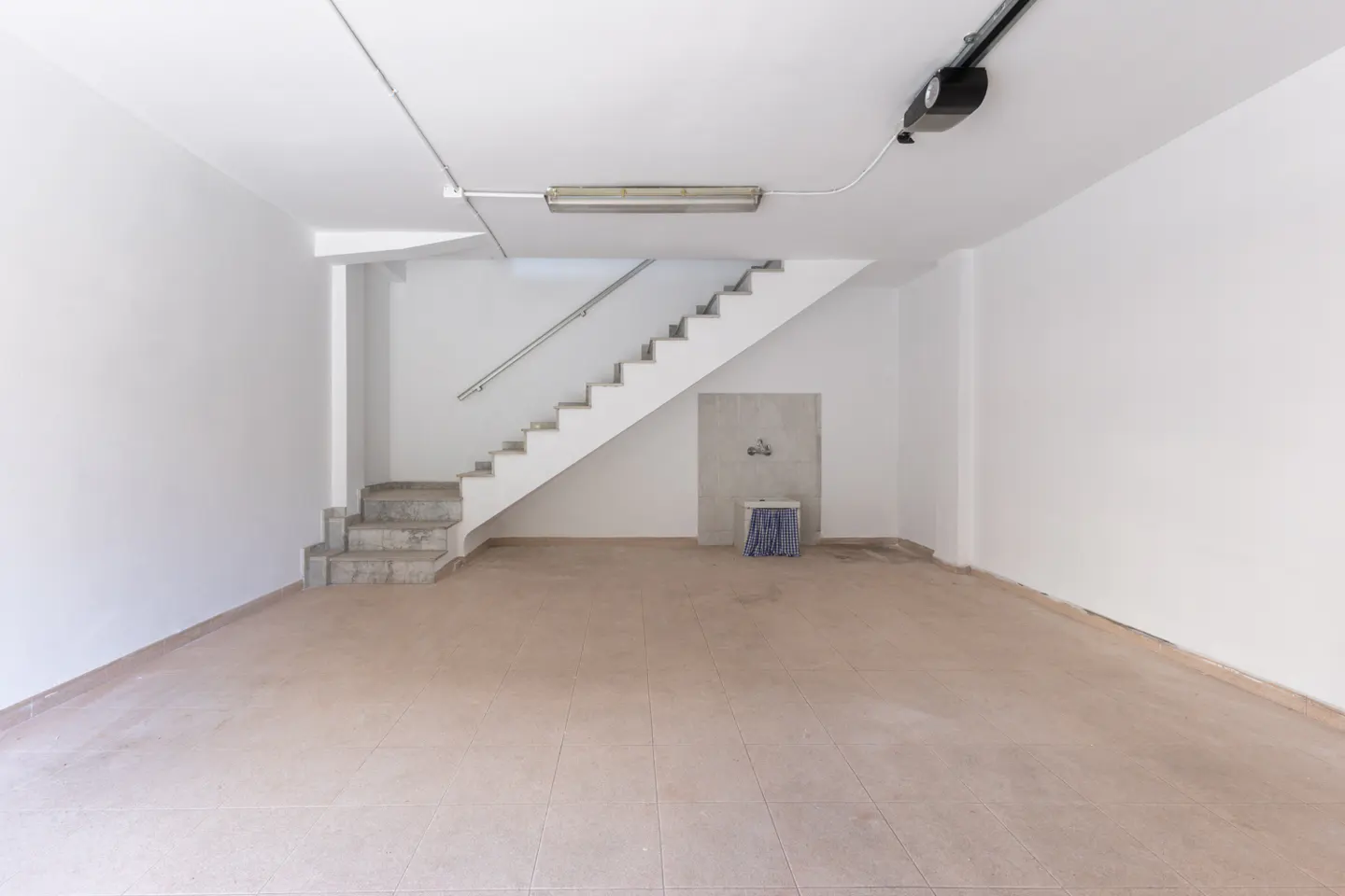 Bright, empty garage with white walls, tiled floor, and stairs leading up. A utility sink and mop are against the back wall.