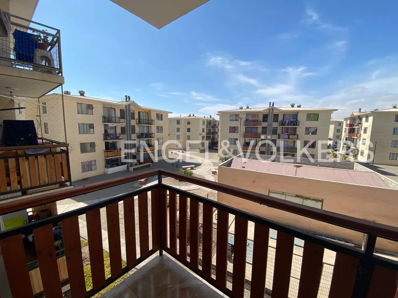 View from a balcony with a brown wood and black metal railing, overlooking apartment buildings under a blue sky.