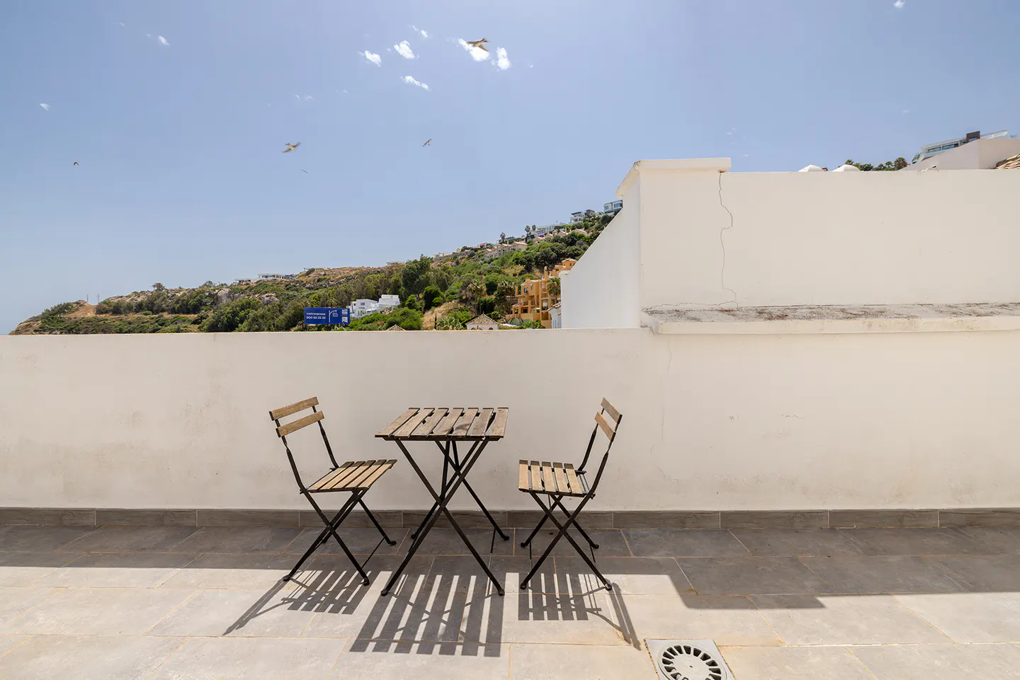 Outdoor patio with a small wooden table and two chairs, white walls, and a view of a green hillside under a blue sky.