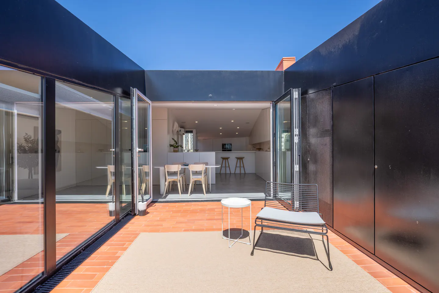 Modern patio with terracotta tiles, a lounge chair, and a small table, opening into a bright, minimalist kitchen interior.