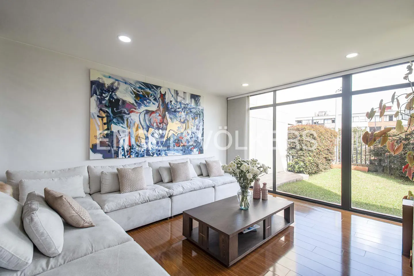 Bright living room with a large abstract horse painting, white sectional sofa, wood floor, and a view of a green garden.