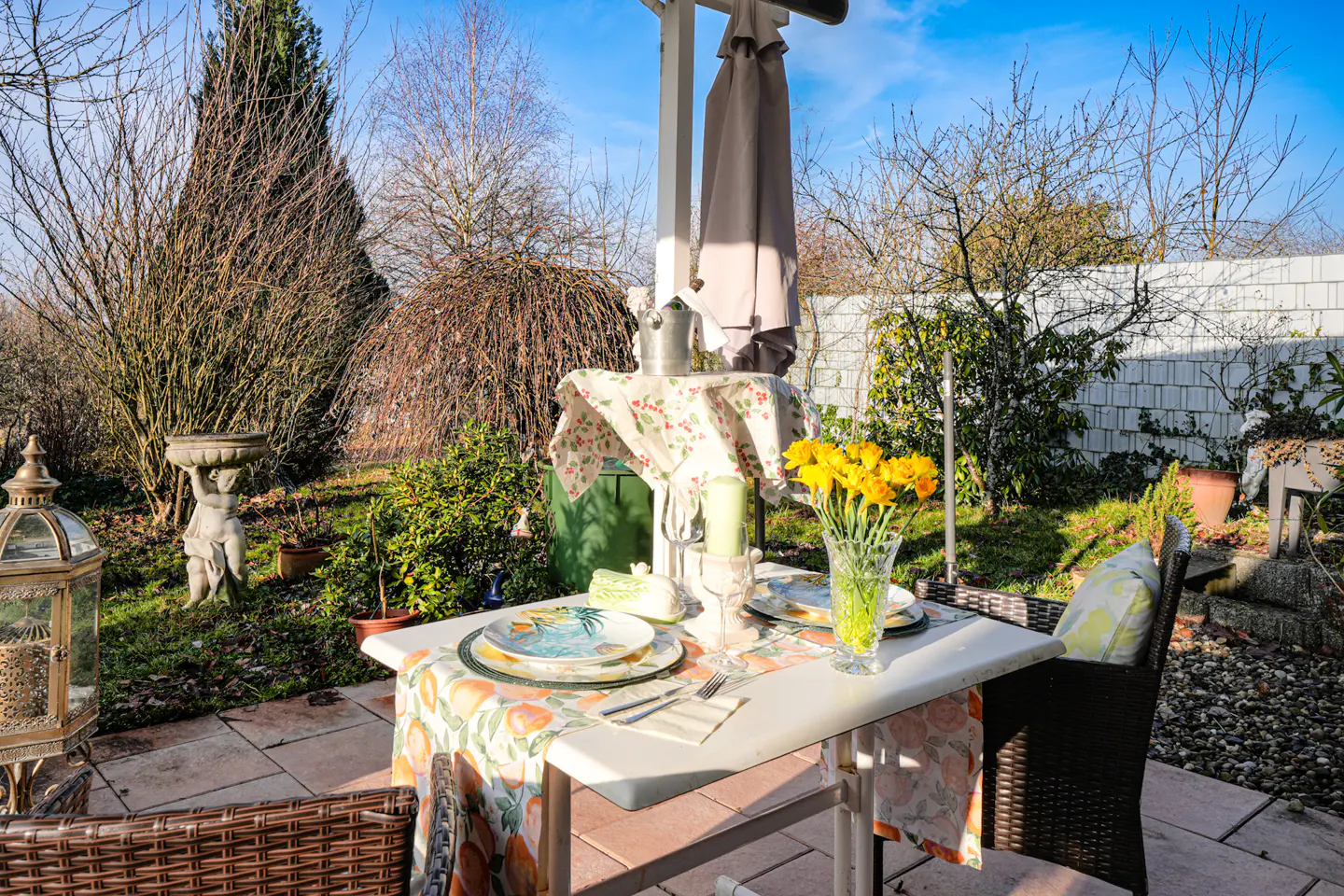 Outdoor patio with a white table set for two, yellow daffodils in a vase, and a garden backdrop.