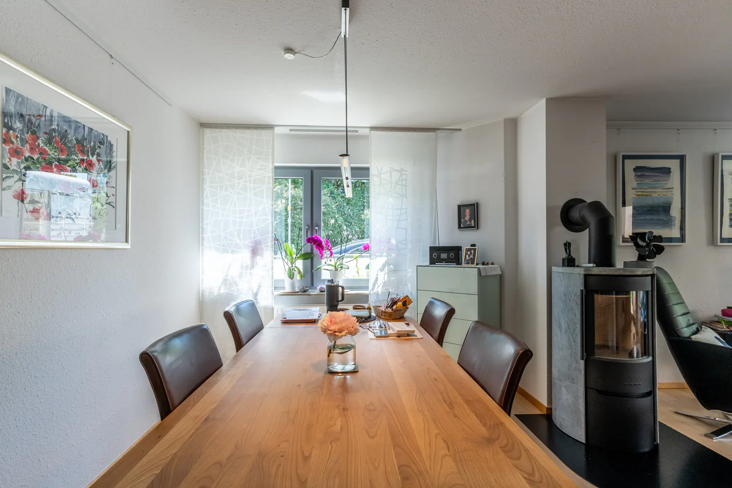 Dining room with a long wooden table, four brown chairs, a modern wood-burning stove, and a window with sheer curtains.