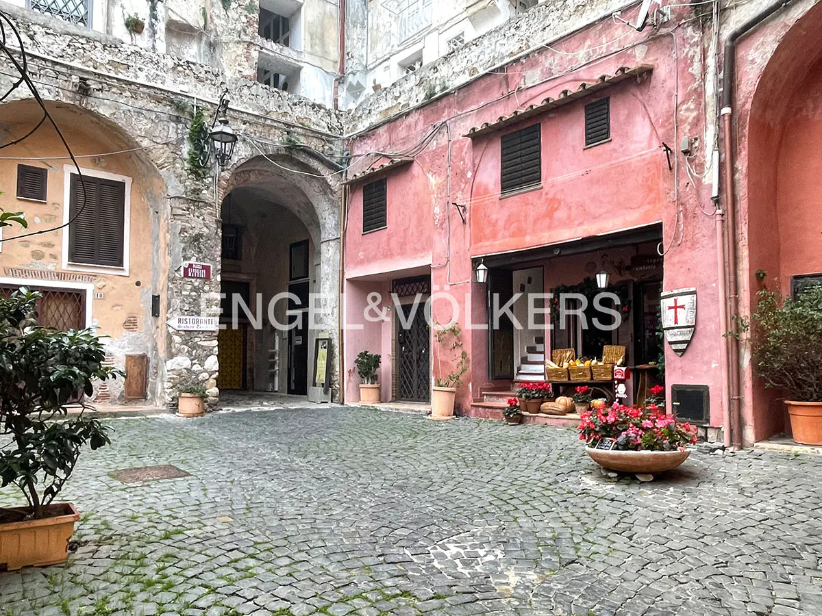 A cobblestone courtyard in Italy featuring a pink building with a shop entrance, potted flowers, and stone archways.