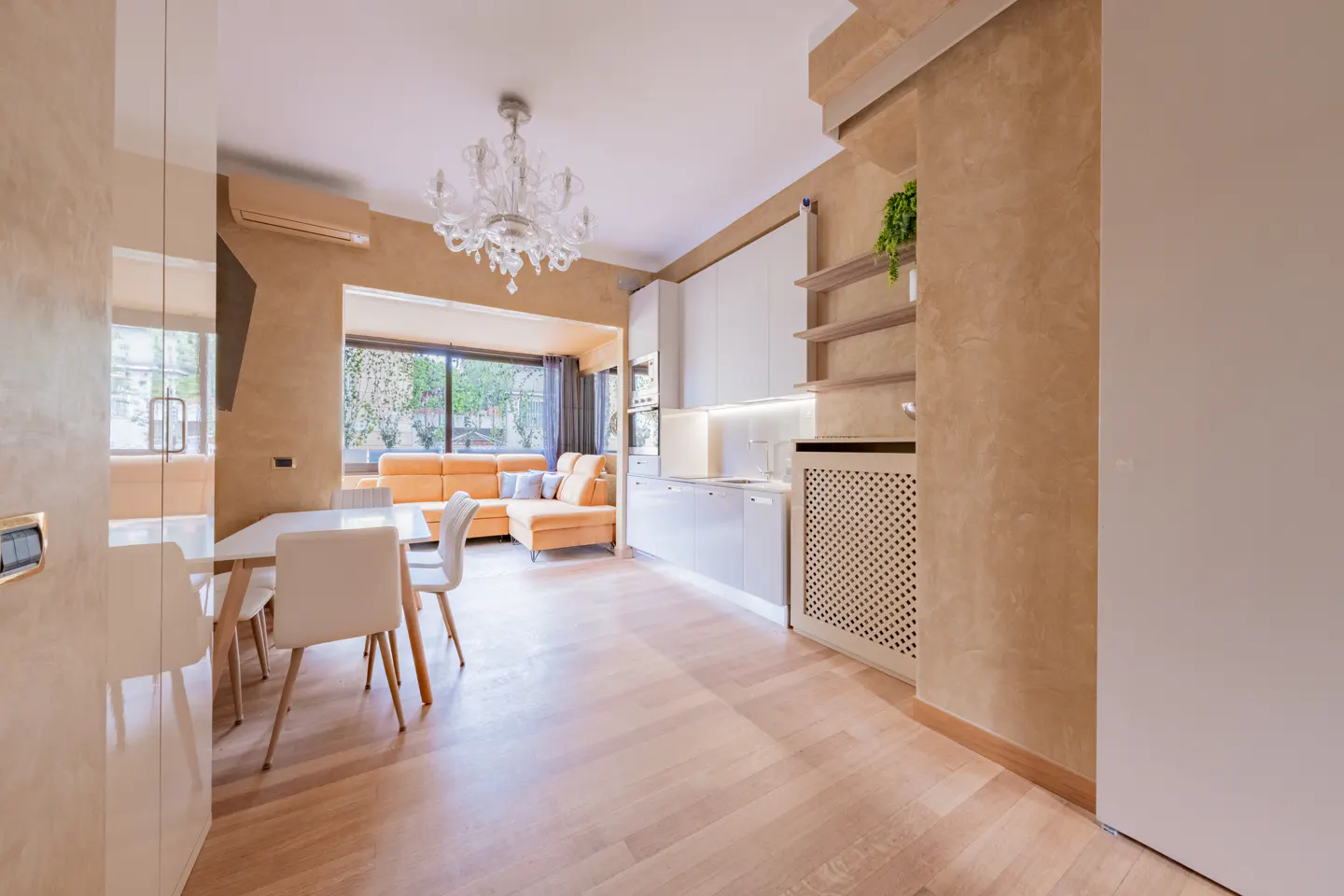 A bright, open-concept apartment with a white kitchen, dining table, and orange sofa. A chandelier hangs above the living area.