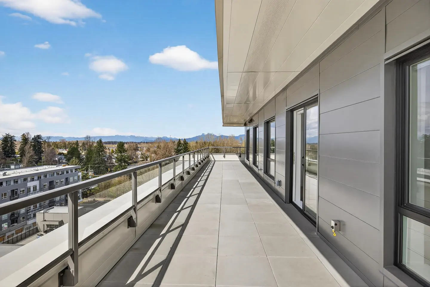 Modern apartment balcony with metal railing, tiled floor, and panoramic views of trees and distant mountains under blue sky.