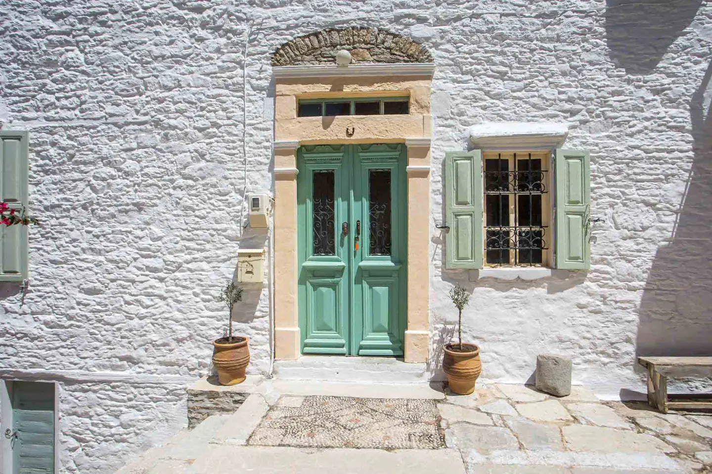 A white stone building with a light green double door, matching shutters, and potted plants on a stone patio.