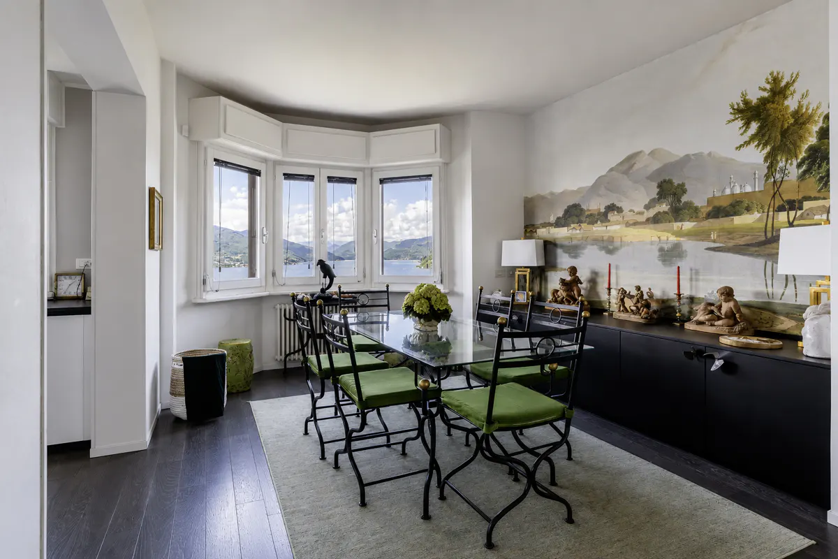 Dining room with black metal chairs with green cushions, glass table, mural, and bay window with lake view.