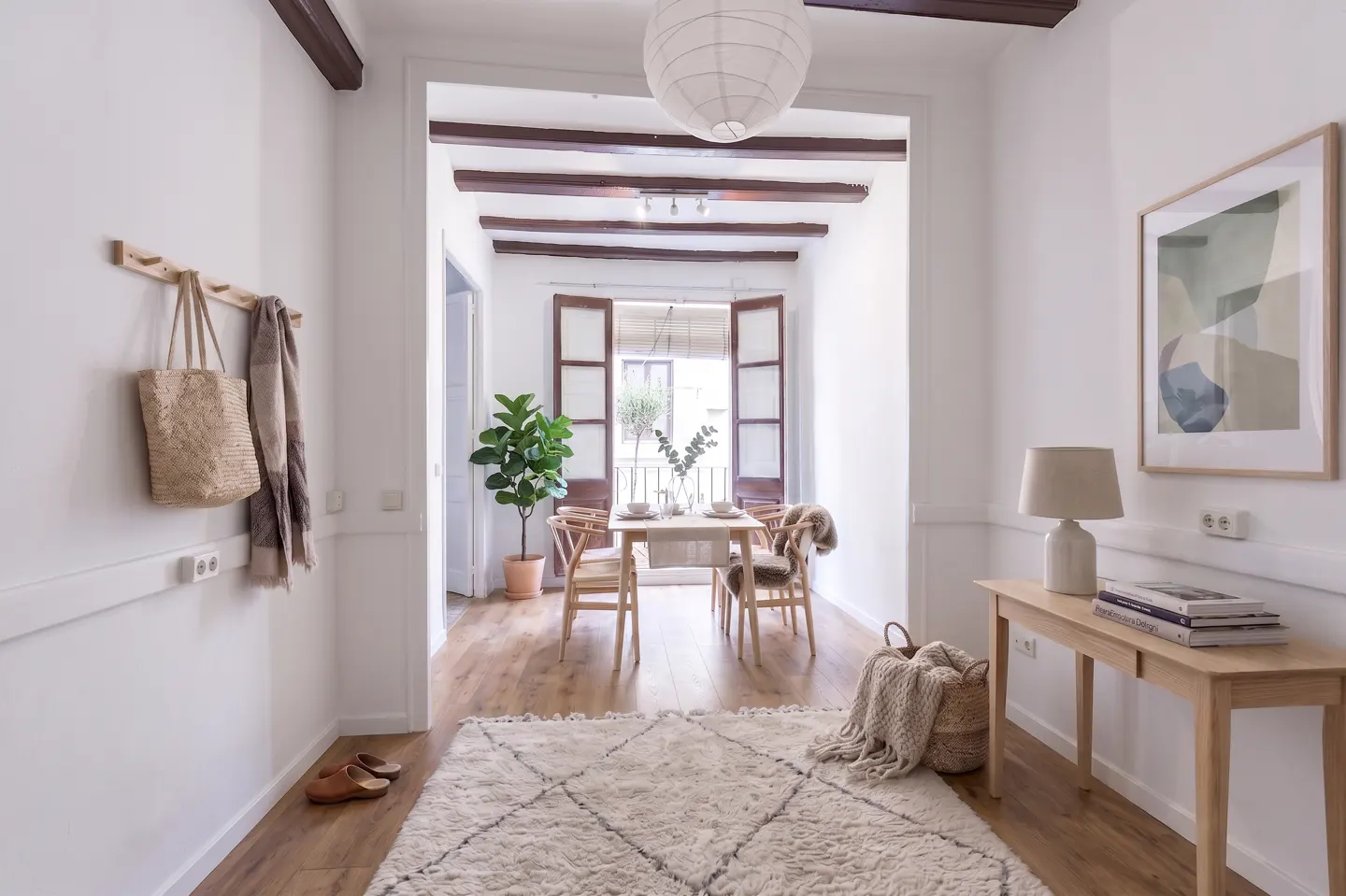 Hallway with white walls, wood floors, and a view into a dining area with a table and chairs. A rug, table, and art decorate the hall.