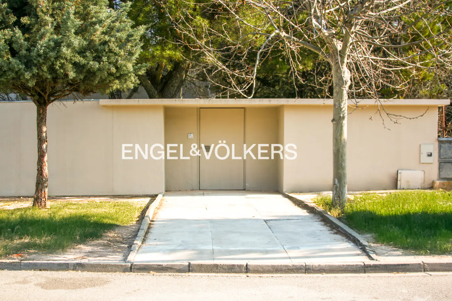 A beige wall with a central door, framed by trees and a concrete walkway leading to the entrance.