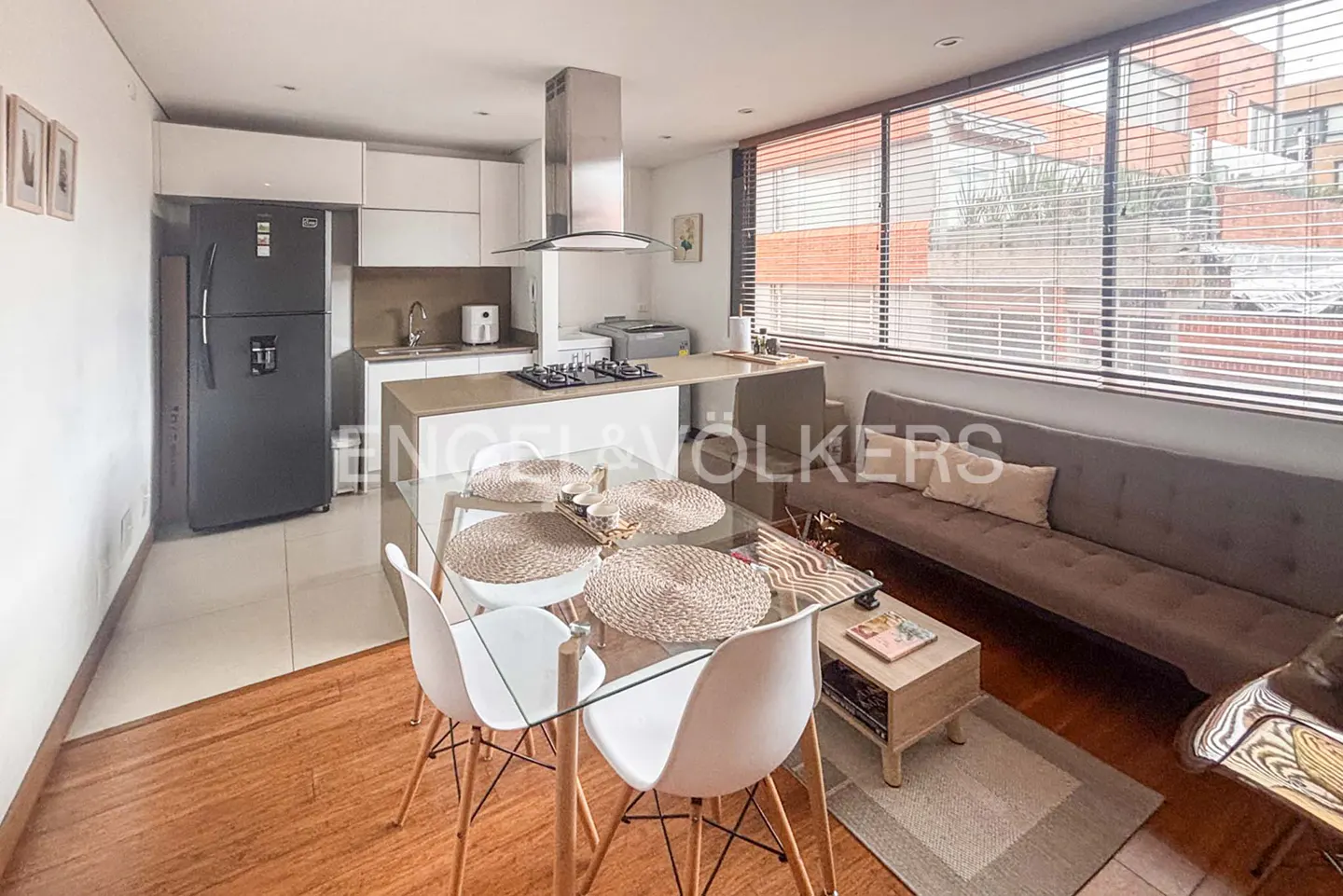 Open-concept apartment with a glass dining table, white chairs, and a gray sofa near a window with blinds. Modern kitchen in the background.