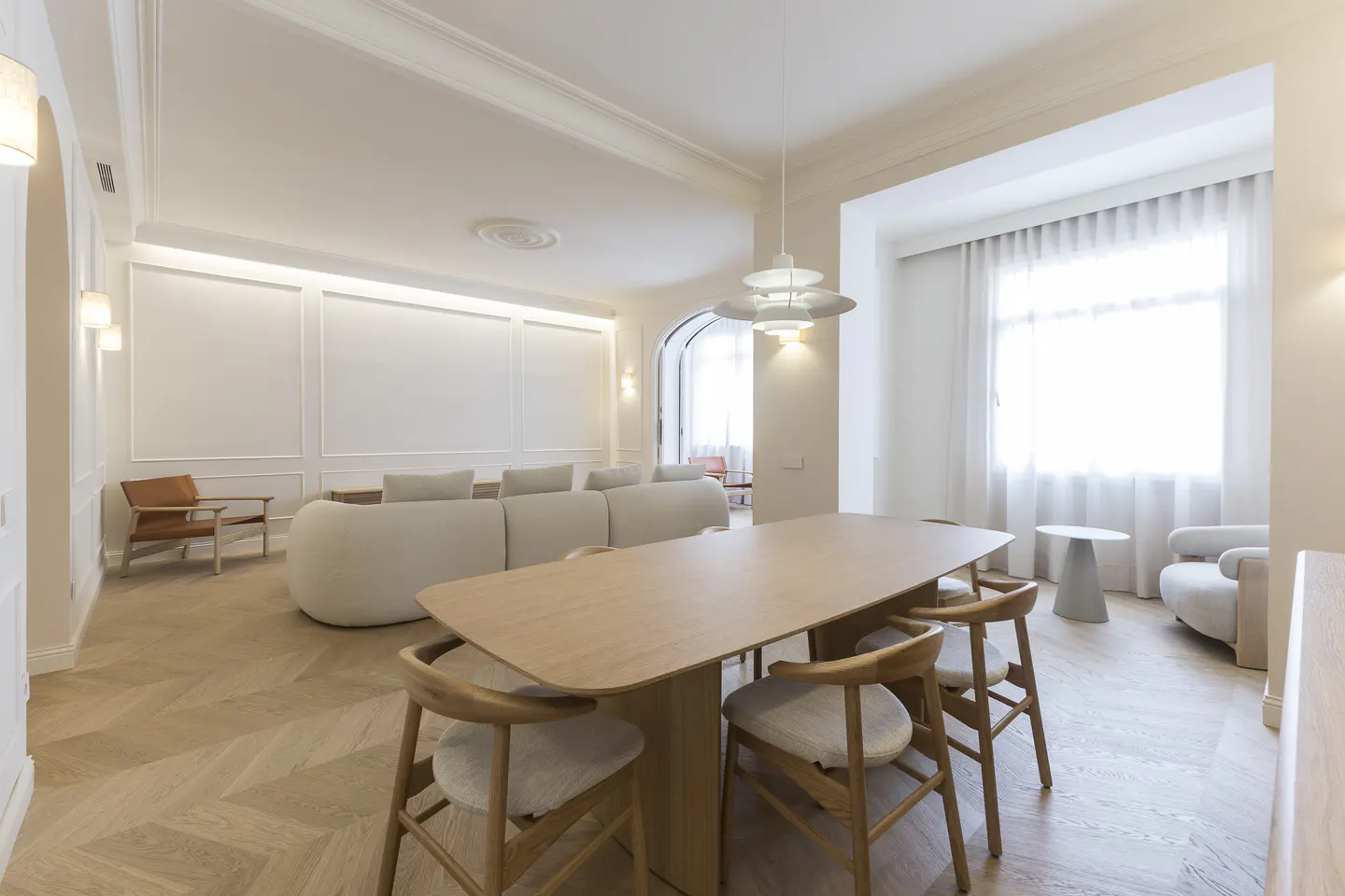 Bright, modern living room with a long wooden table and chairs in the foreground, and a white sofa in the background.
