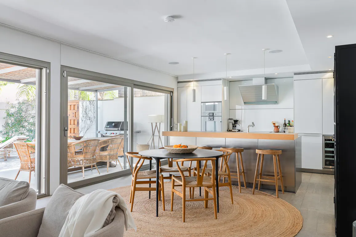 Bright, modern kitchen with a round table, wooden chairs, and a jute rug. Sliding glass doors lead to an outdoor patio with wicker furniture.