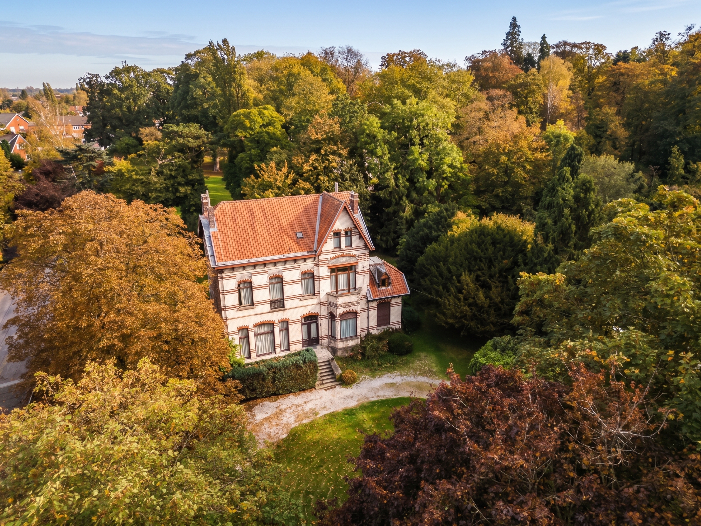 Aerial view of a classic brick house with a red tile roof, nestled in a lush, autumn-colored forest with a gravel driveway. [AI-generated text]