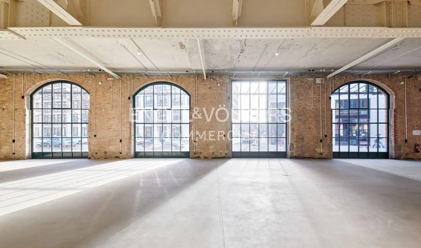 Empty industrial space with brick walls, concrete floor, and arched windows. Sunlight streams through the windows.