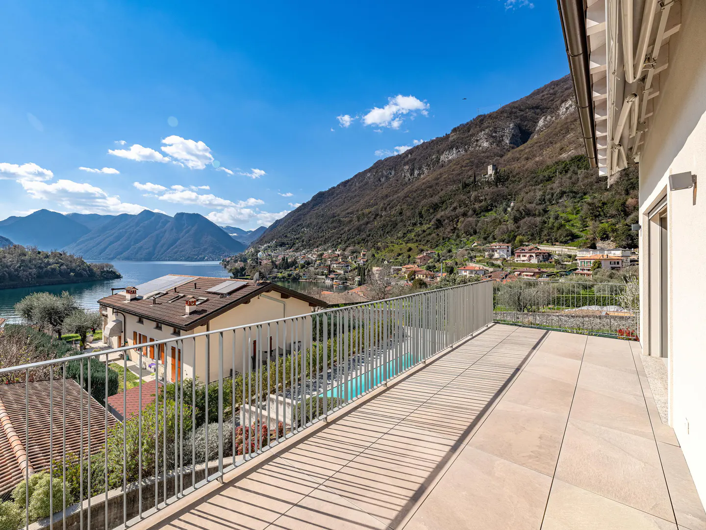 Balcony view of a lake and mountains. Gray railings line the tiled balcony. Houses dot the hillside. Blue sky with clouds.