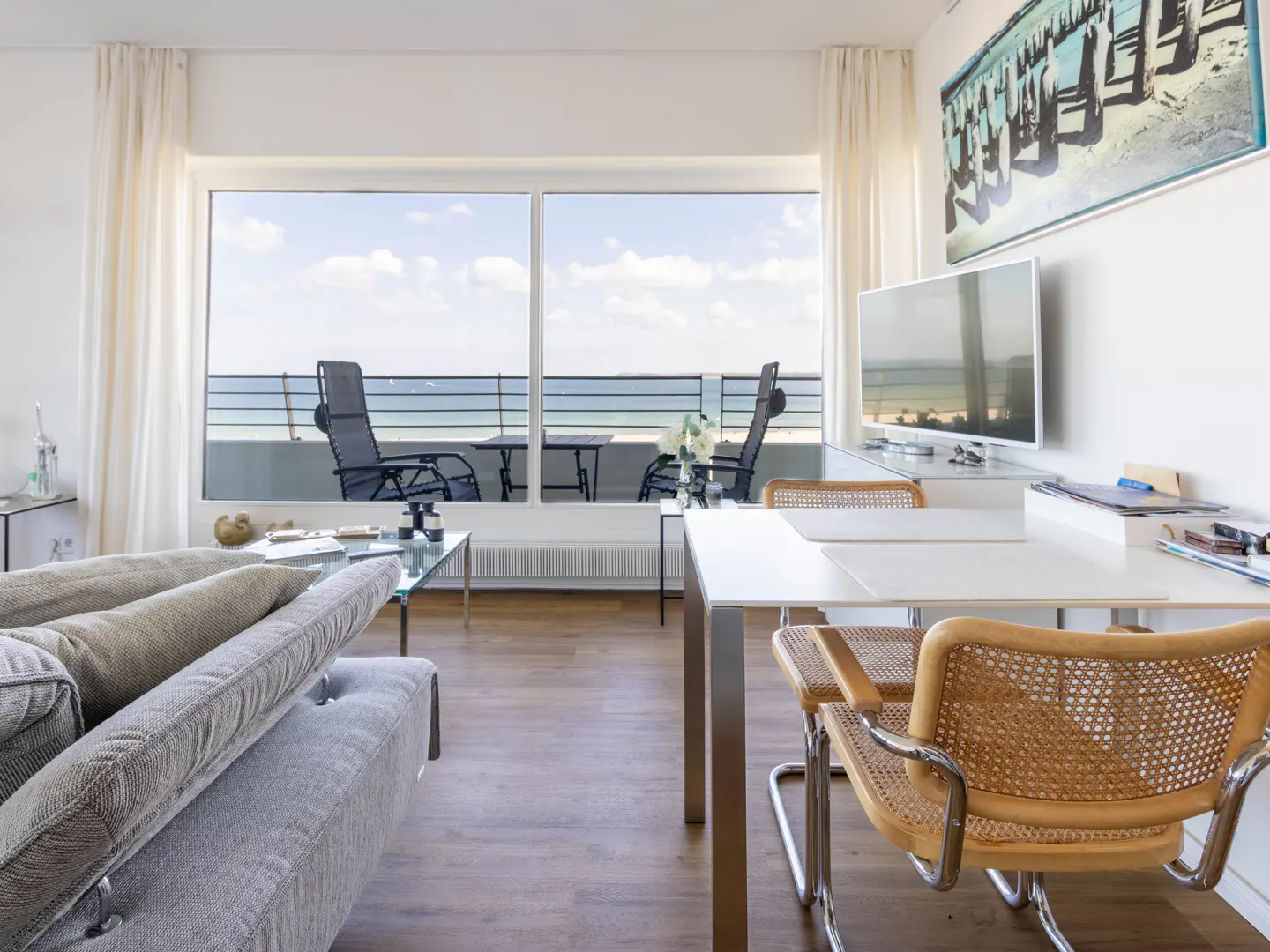 Bright living room with a gray sofa, white walls, and a large window overlooking the ocean. A white table and wicker chairs are in the foreground.
