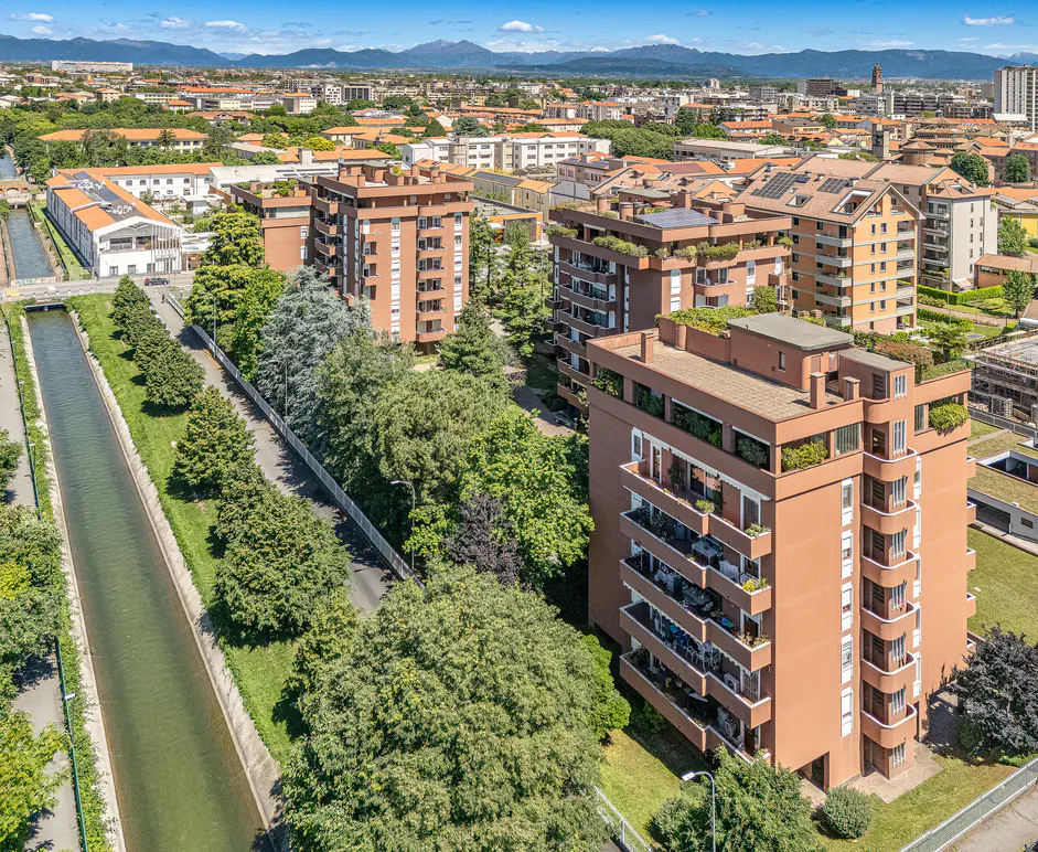 Aerial view of terracotta apartment buildings with balconies, green trees, and a canal in a city with mountains in the background.