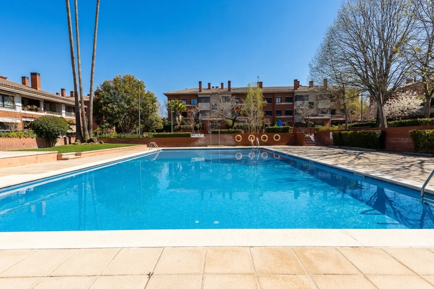 Outdoor pool with blue water, surrounded by a tiled deck. Apartment buildings and trees are in the background under a clear blue sky.