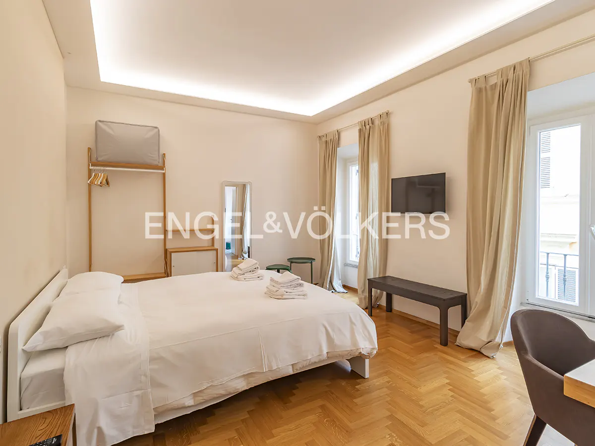 Bright bedroom with white linens, wood floors, and beige curtains. A TV hangs on the wall, and a clothing rack stands in the corner.