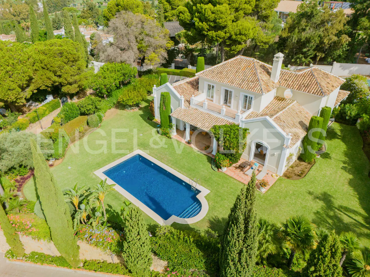 Aerial view of a two-story, beige house with a red tile roof, a blue pool, and green lawn surrounded by trees.