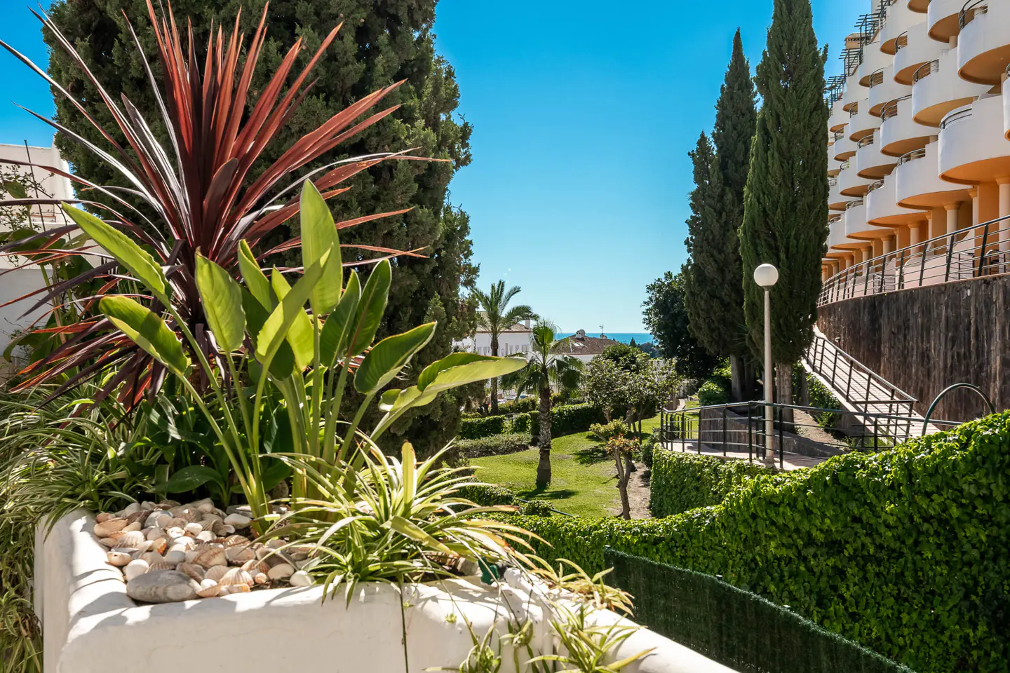 A view of a lush garden with a building in the background on a sunny day. The garden has green plants and trees, and a white planter with rocks.