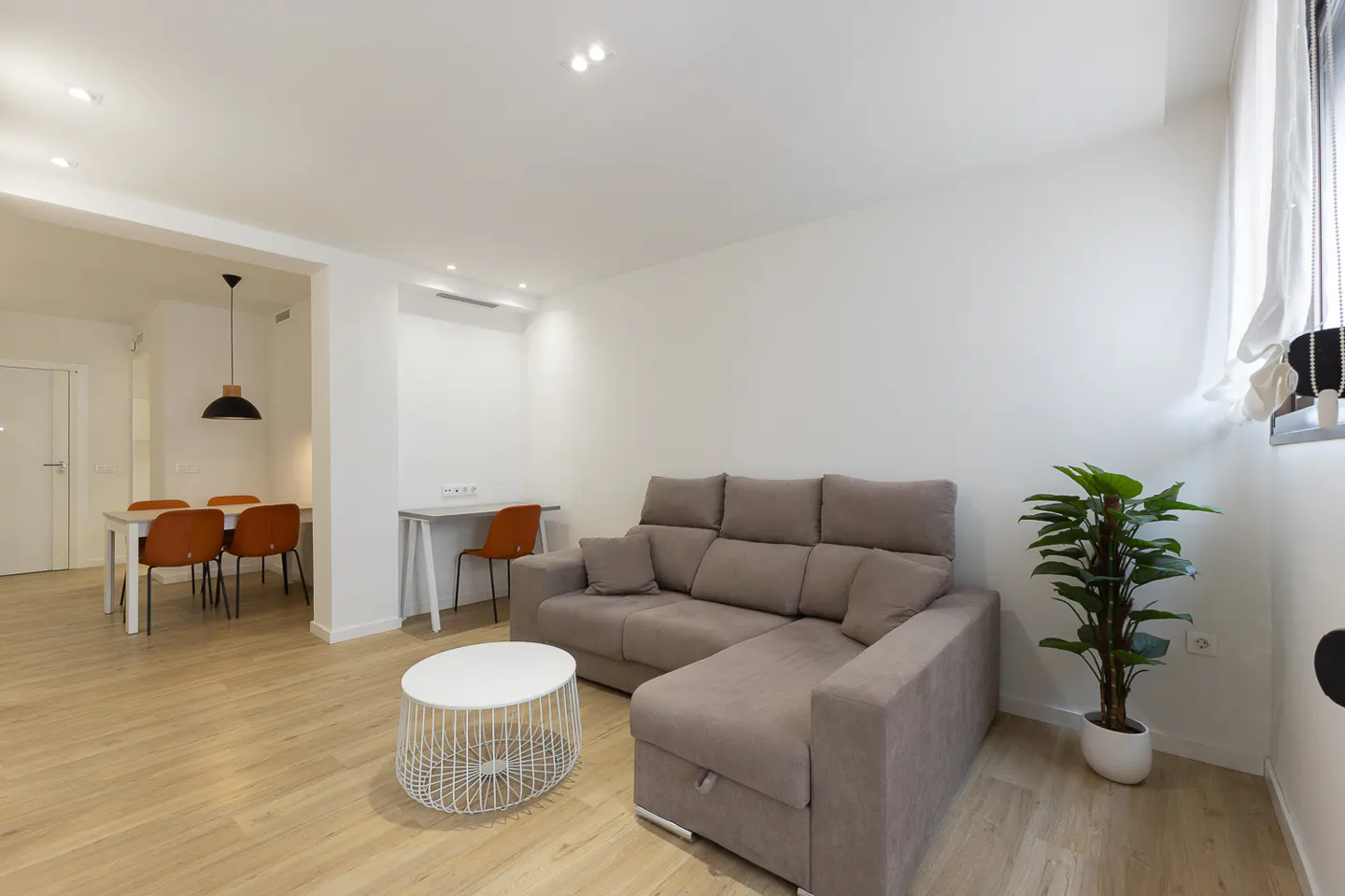 Modern living room with a grey sectional sofa, white wire coffee table, potted plant, and a dining area in the background.