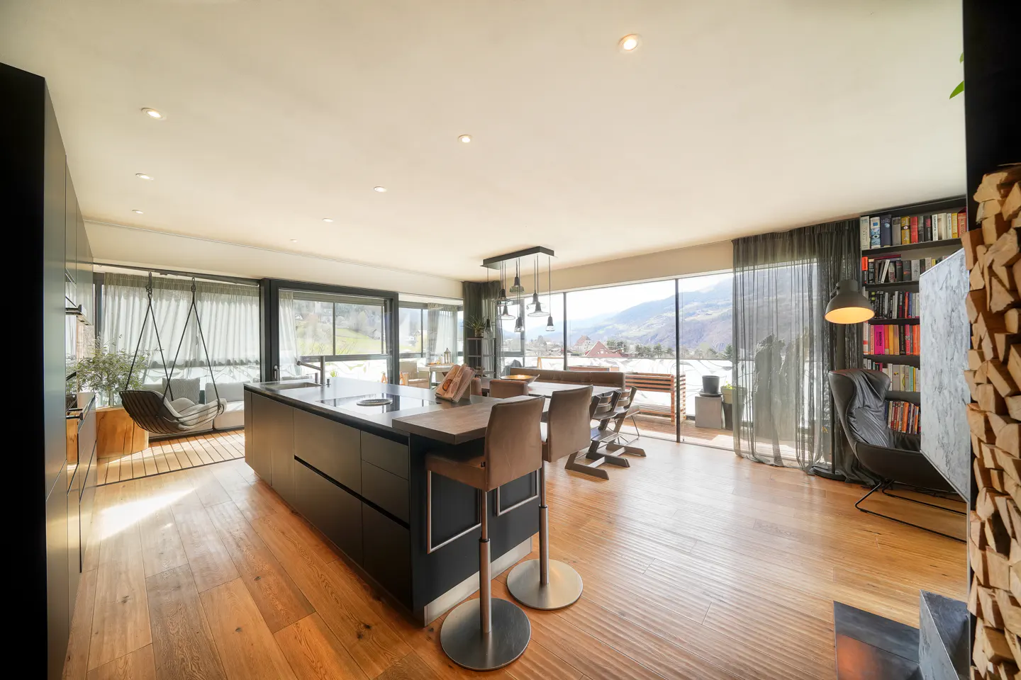 Bright, open-concept living space with a black kitchen island, bar stools, and a dining area. Large windows offer mountain views. Bookshelves and a hanging chair add character.