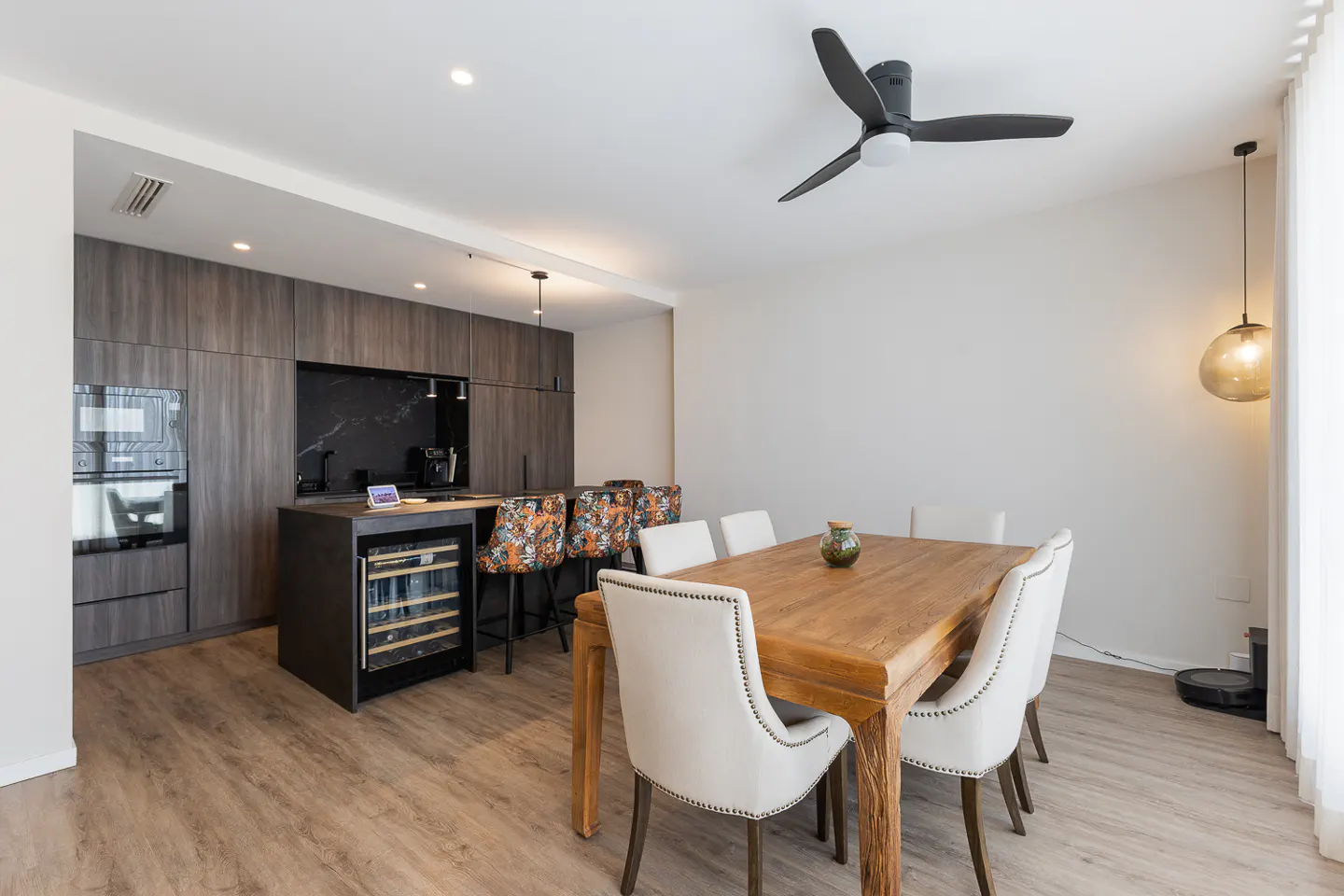 Open-concept kitchen and dining area with wood floors, a black ceiling fan, and a wooden table with white chairs.