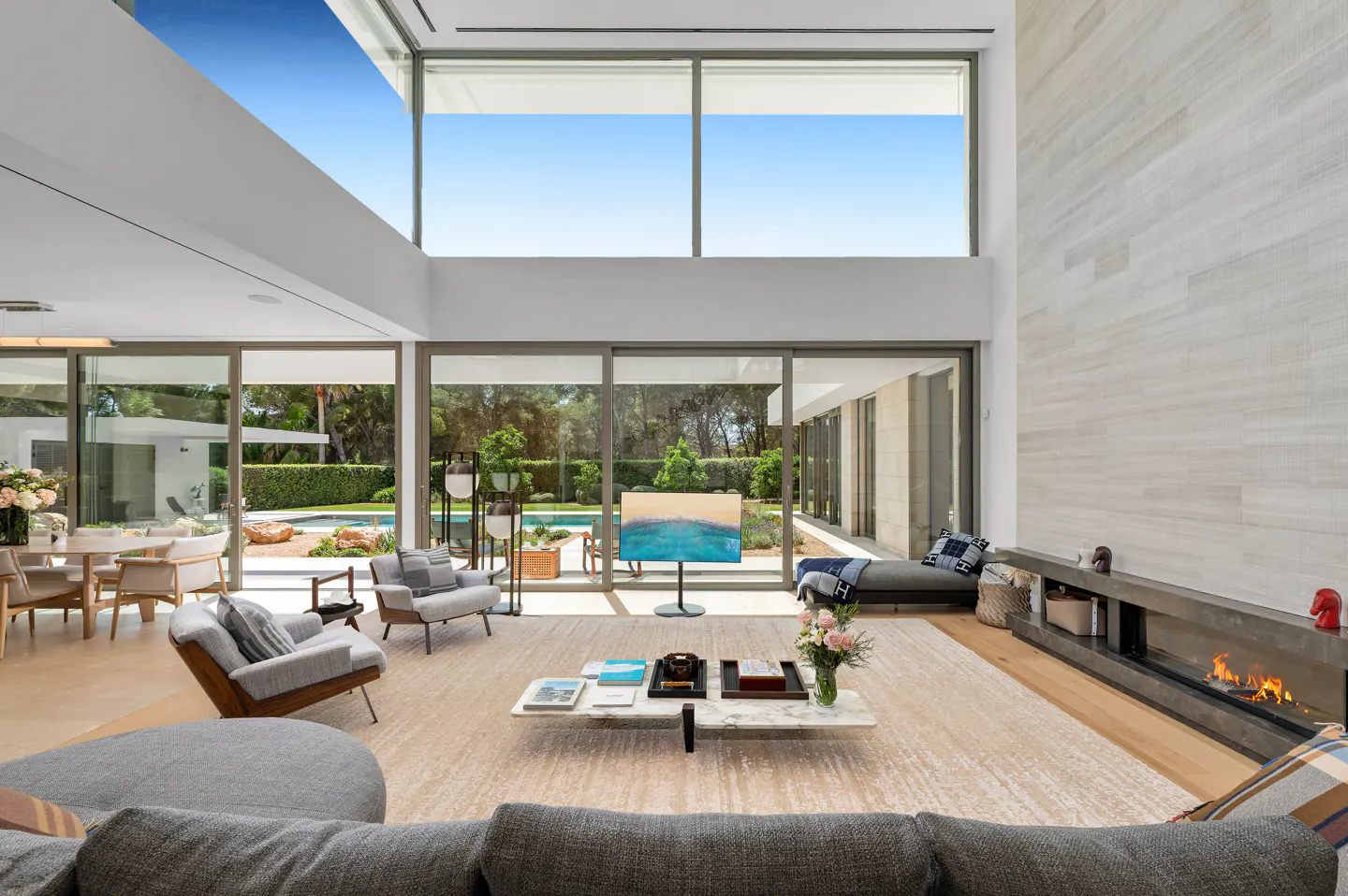 Bright, modern living room with gray furniture, a fireplace, and large windows overlooking a pool and garden.