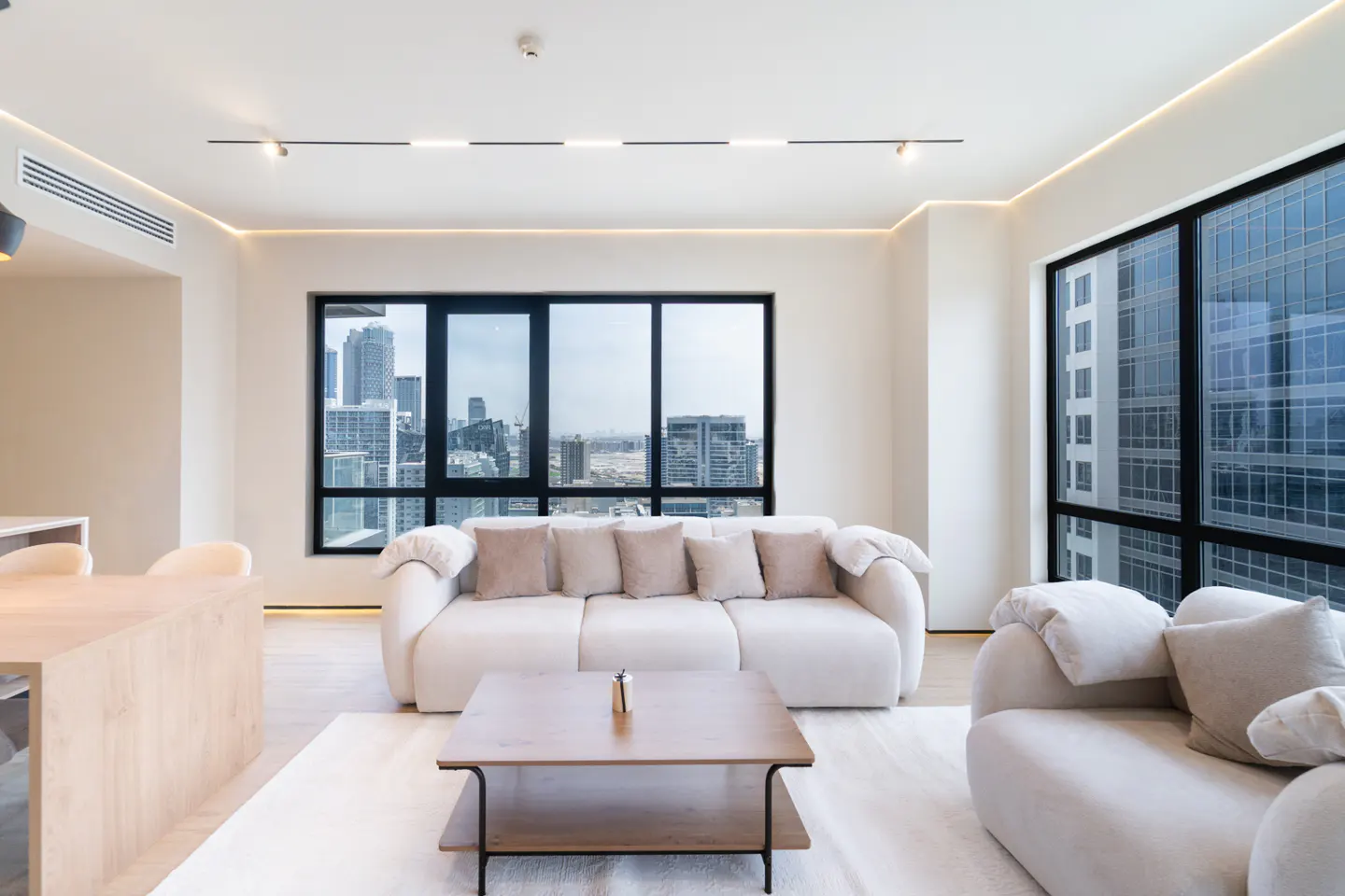 Bright living room with a white sofa, wood table, and city views through large windows.
