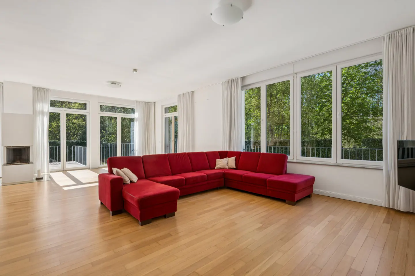 Bright living room with hardwood floors, white walls, and large windows. A red sectional sofa sits in the center. Fireplace and balcony visible.