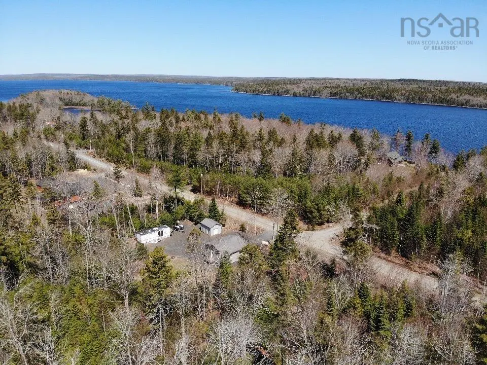 Aerial view of a wooded property with a house, trailer, and outbuilding near a blue lake under a clear sky.