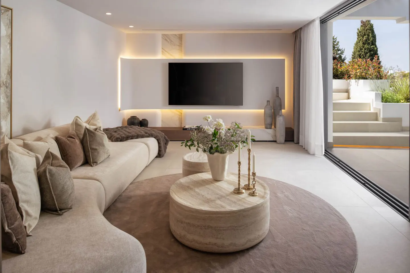 Bright living room with a beige curved sofa, round rug and coffee table, and a TV with LED lighting. Sliding glass doors lead to an outdoor patio.