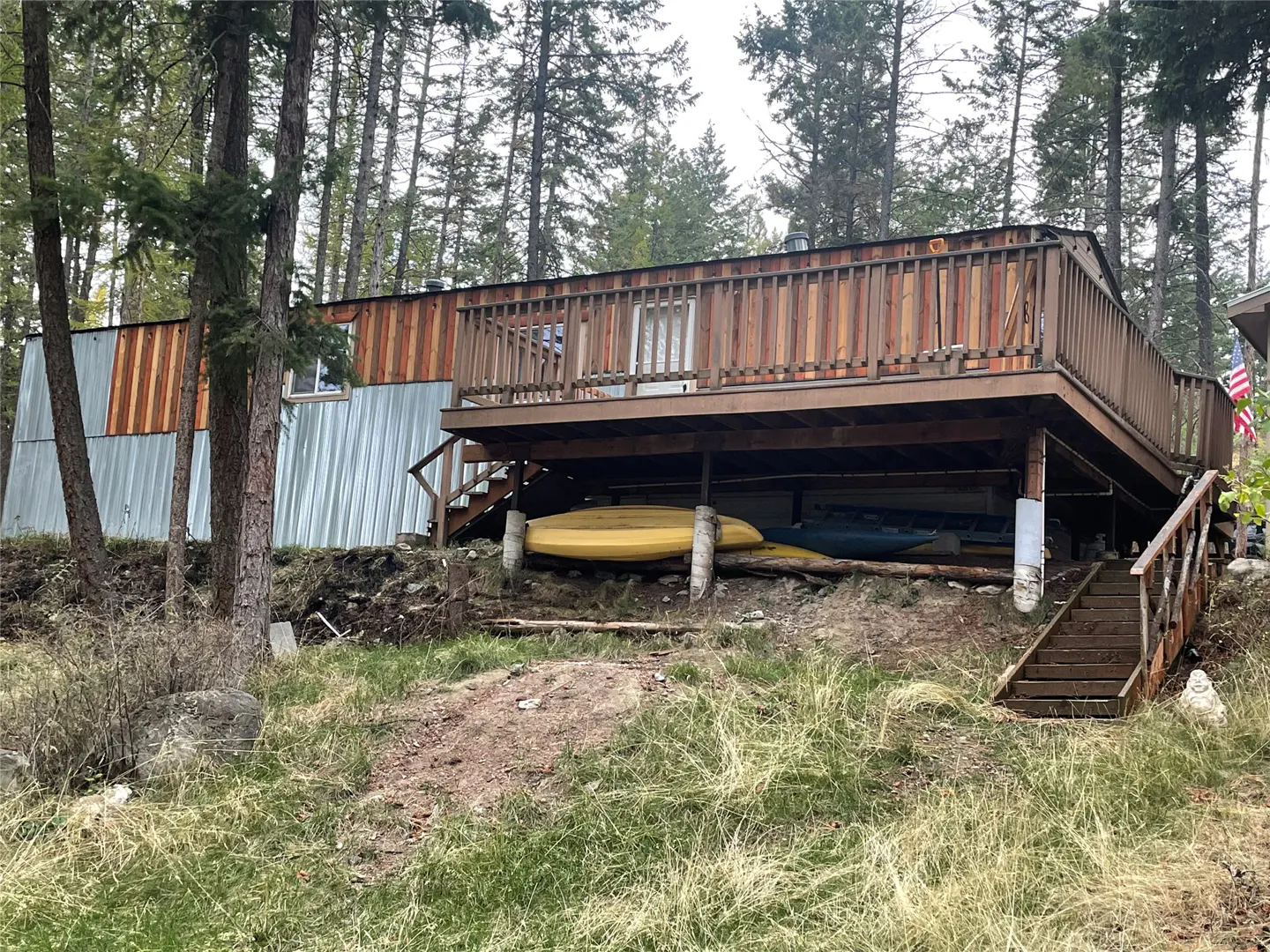 Exterior view of a house with a wooden deck, stairs, and kayaks stored underneath, surrounded by trees.