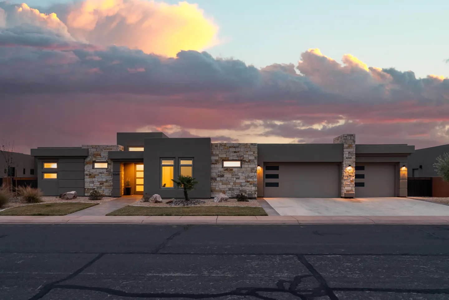 Modern, gray, single-story home with stone accents and a two-car garage under a colorful sunset sky.