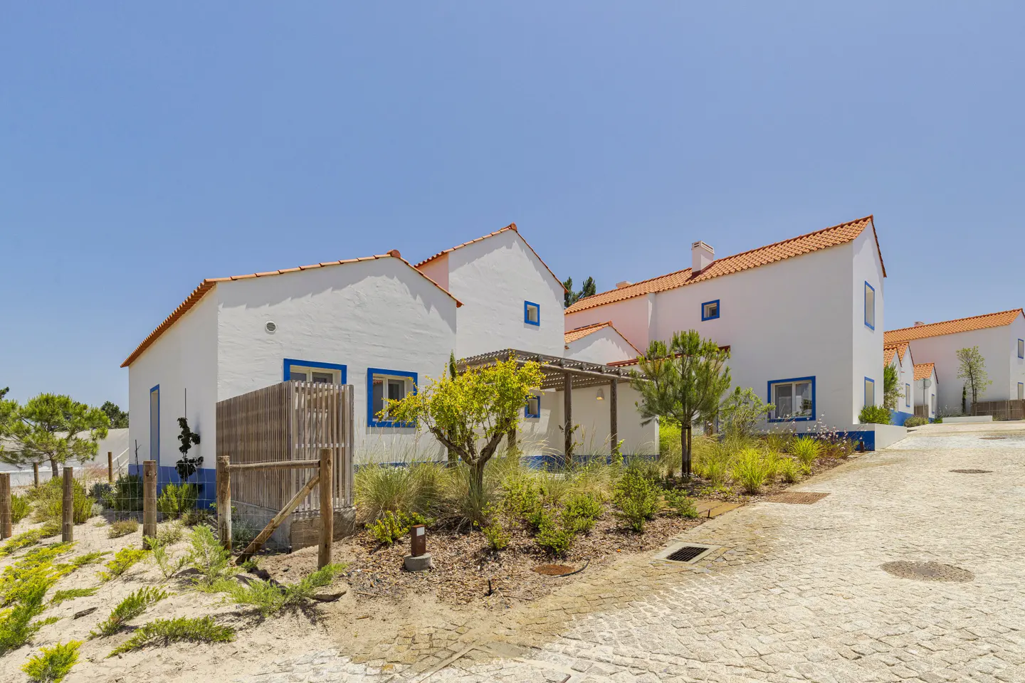 White villas with orange roofs and blue trim set against a clear blue sky, featuring a cobblestone path and landscaping.