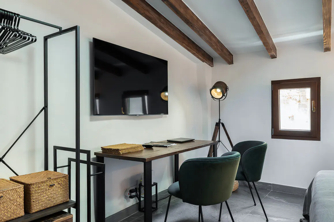 A modern room with a desk, two green chairs, a TV, and a floor lamp. Wicker baskets sit on a shelf. The ceiling has wooden beams.