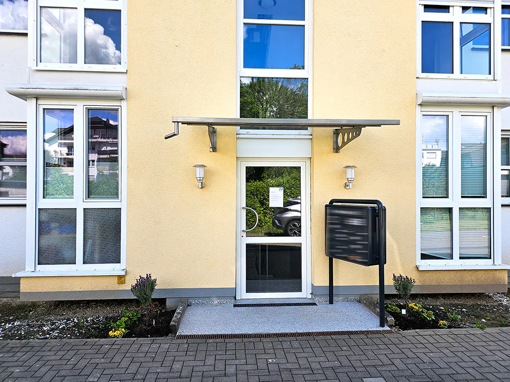Apartment building entrance with a white door, metal awning, and black mailbox. The building is yellow with white-framed windows.