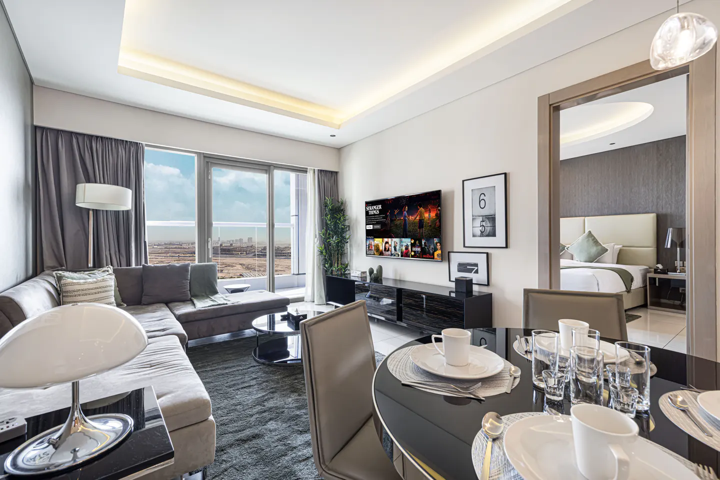 Modern apartment interior with gray sectional sofa, black table, and TV showing "Stranger Things". Dining table set for a meal. Bedroom visible through doorway.