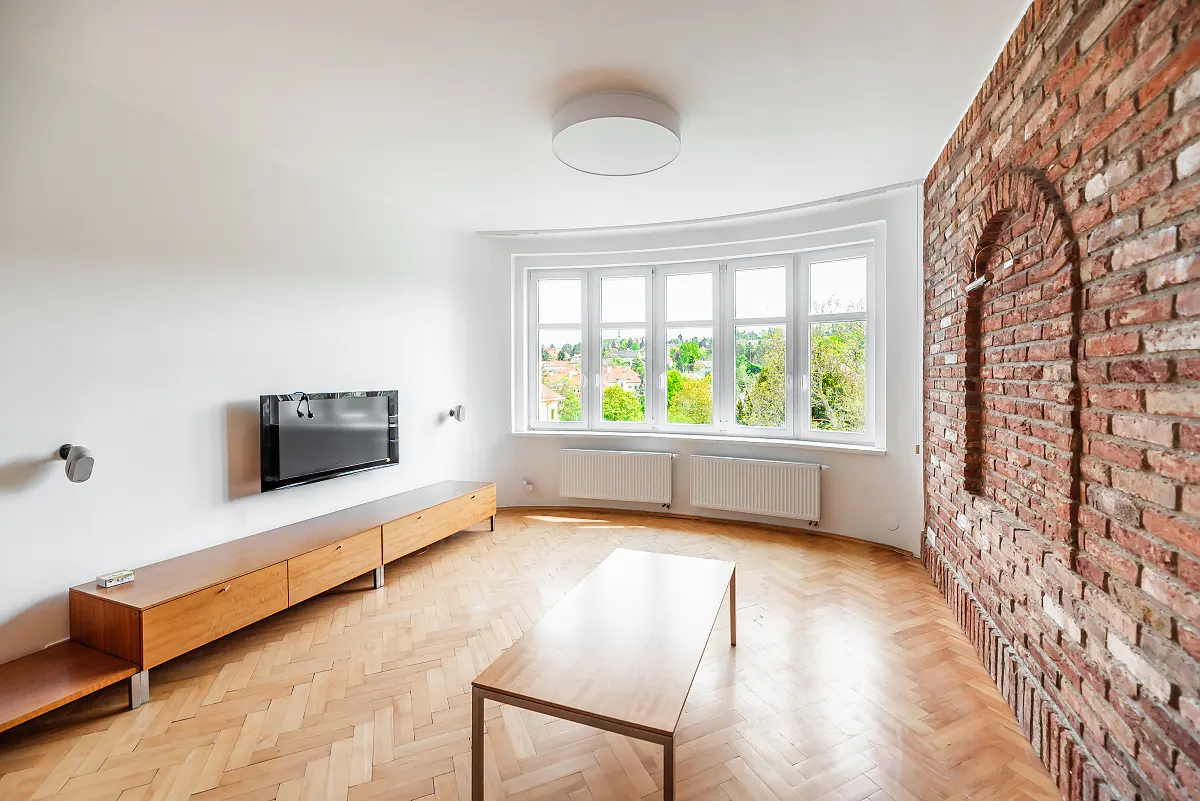 Bright living room with herringbone wood floors, white walls, and a brick accent wall. A large window overlooks green trees.