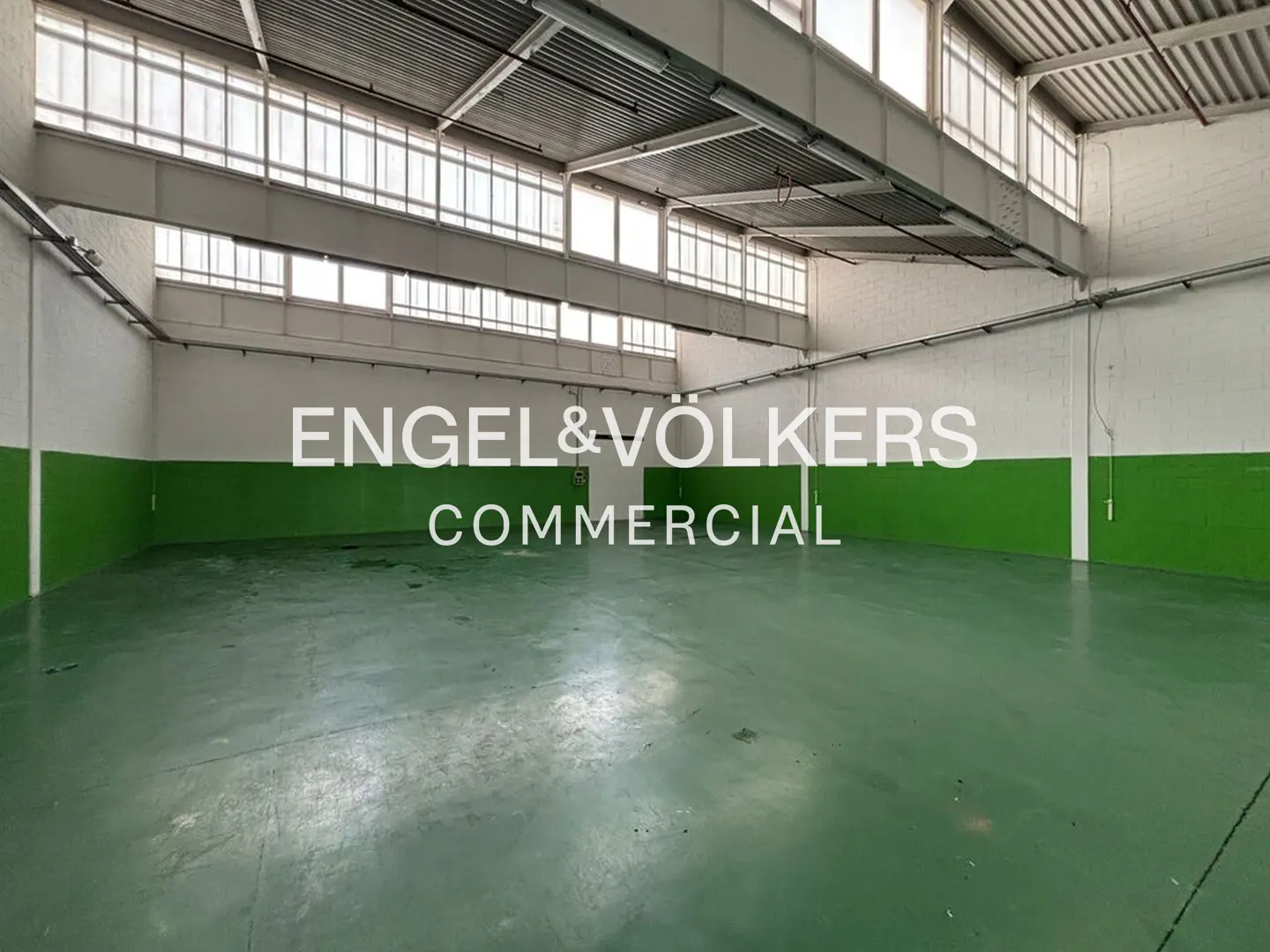 Empty commercial space with green floor and white walls. Windows line the top of the walls. Engel & Völkers logo is superimposed.