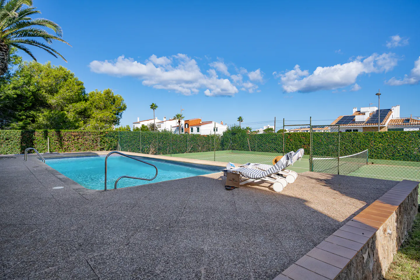 Outdoor pool with metal railings, a striped lounge chair, and a tennis court with a net, all under a blue sky.