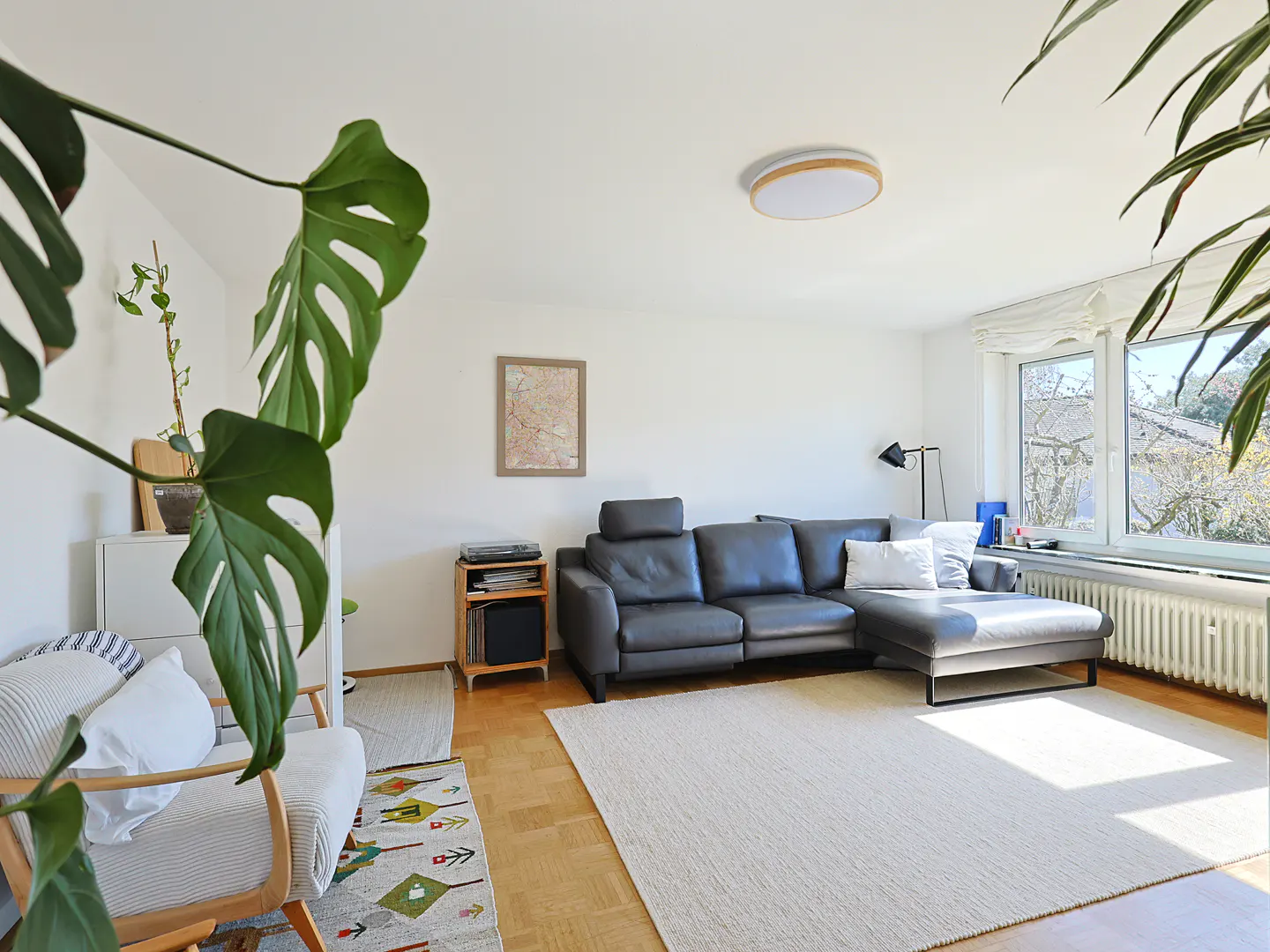 Bright living room with a gray leather sectional sofa, white rug, and large windows. Plants add a touch of nature.